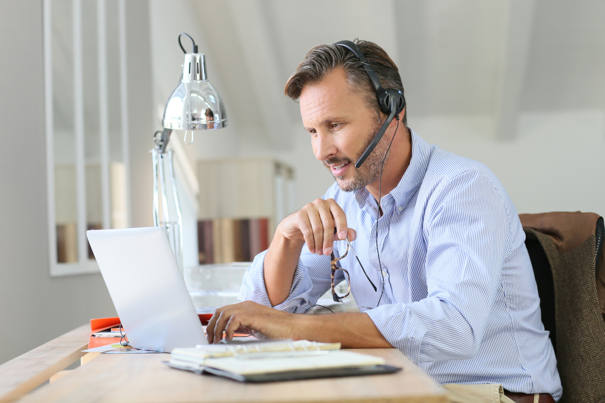 An older man using a headset while working on his laptop at his desk inside his home