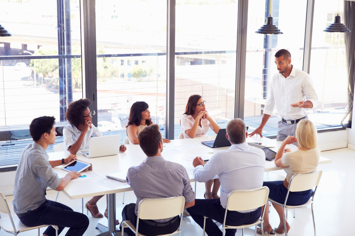 Group of Seven Business People Seated Around Meeting Table Looking at with Man in White Dress Shirt Standing in Front of Room